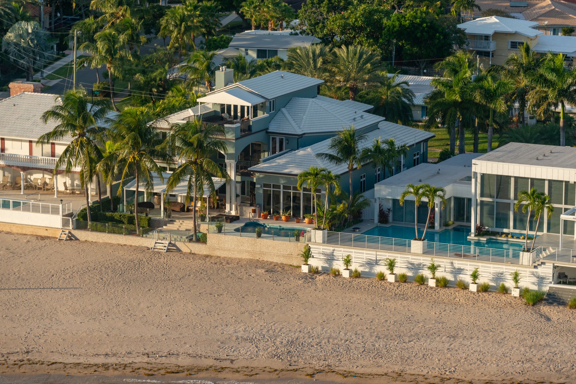 Aerial view of luxury beachfront houses with palm trees, pool, and sandy beach.