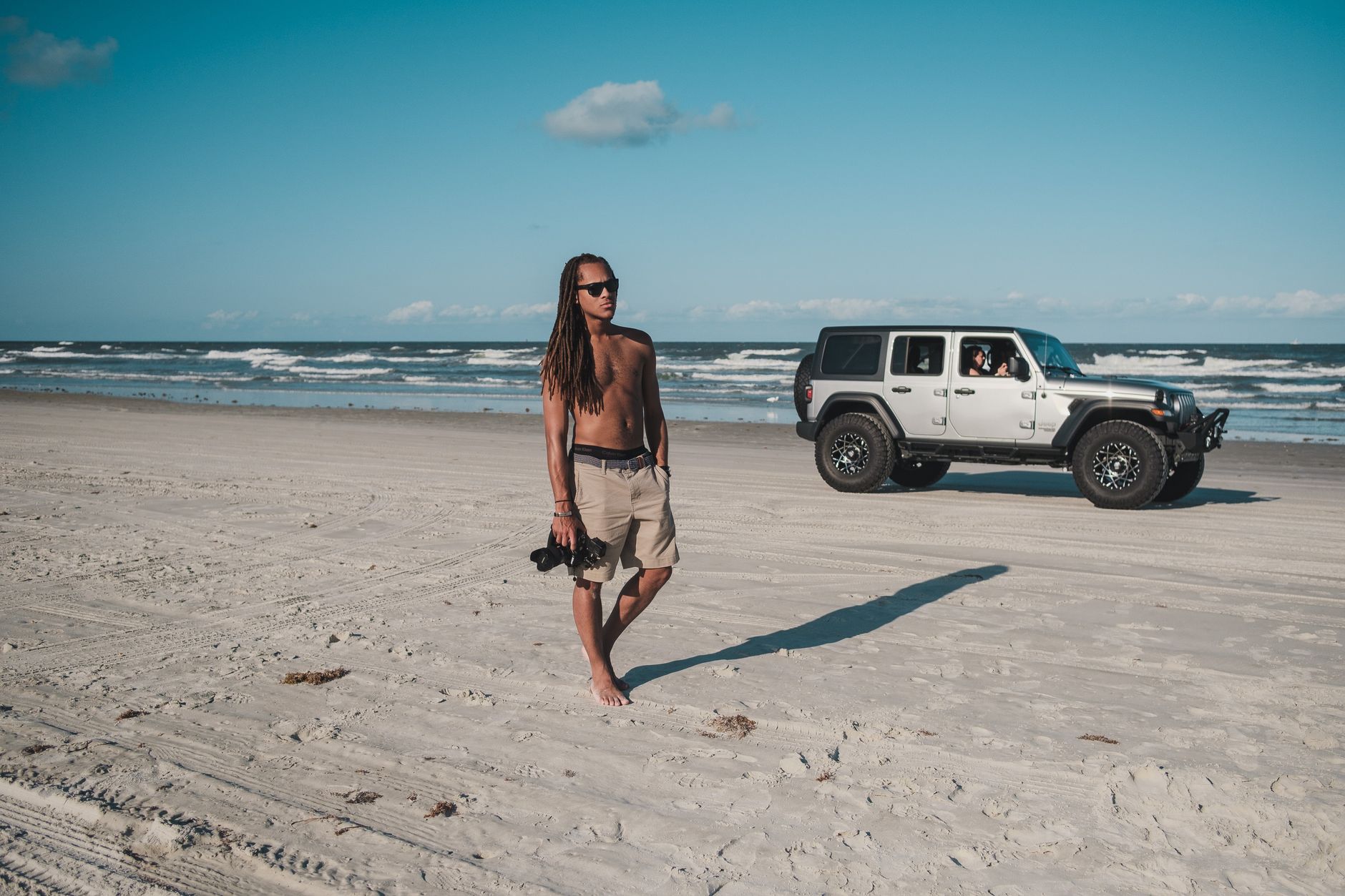 Shirtless man with dreadlocks walking on New Smyrna Beach, Florida near an SUV under a clear blue sky.